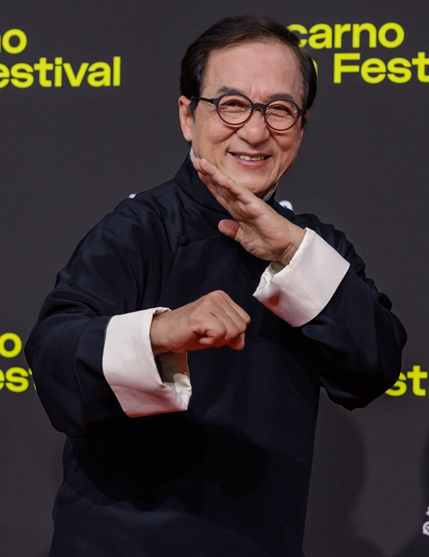 Jackie Chan in a dark Chinese-style jacket and round glasses, smiling and striking a playful martial arts pose on a red carpet at the Locarno Film Festival in 2025